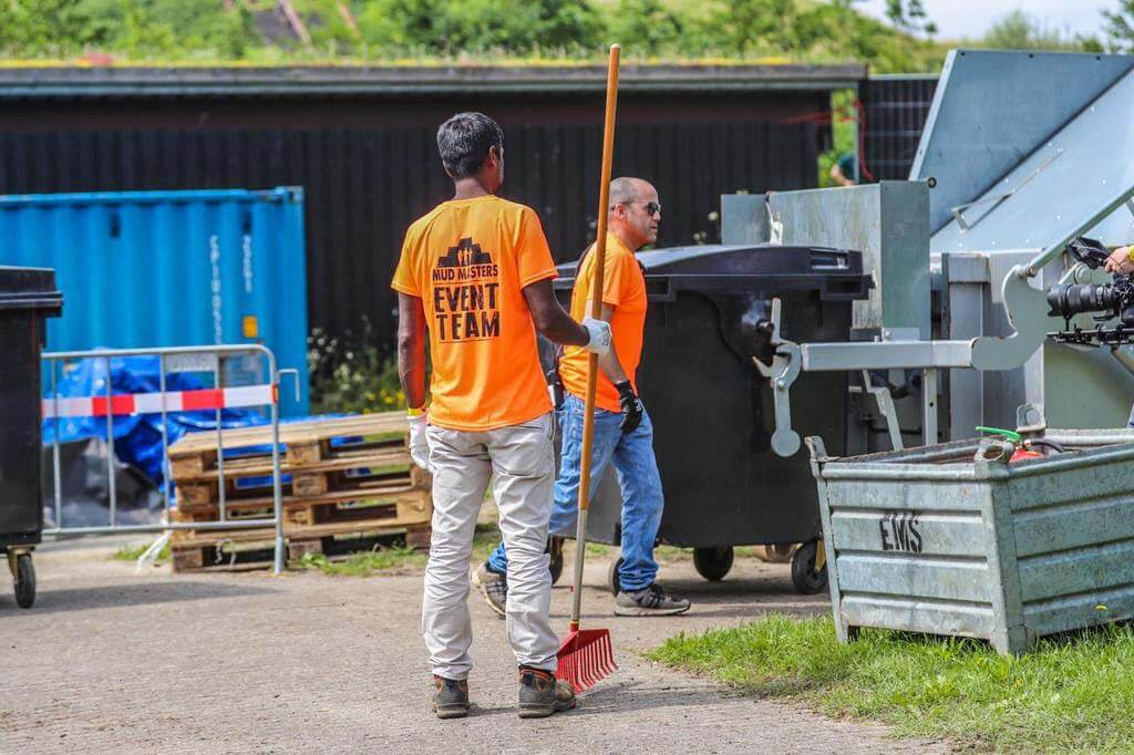 Twee medewerkers in oranje eventteam-shirts die eindschoonmaak uitvoeren bij evenemententerrein, zichtbaar afvalbeheer door What A Waste.