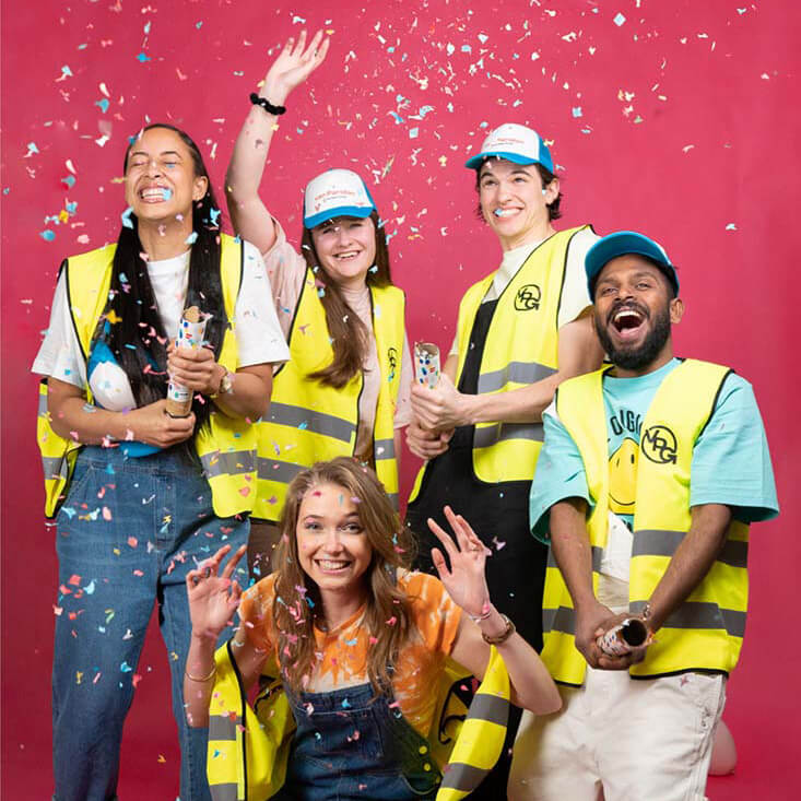 Groepsfoto van het What A Waste team met vijf enthousiaste medewerkers, gekleed in kleurrijke werkkleding, lachend met confetti.