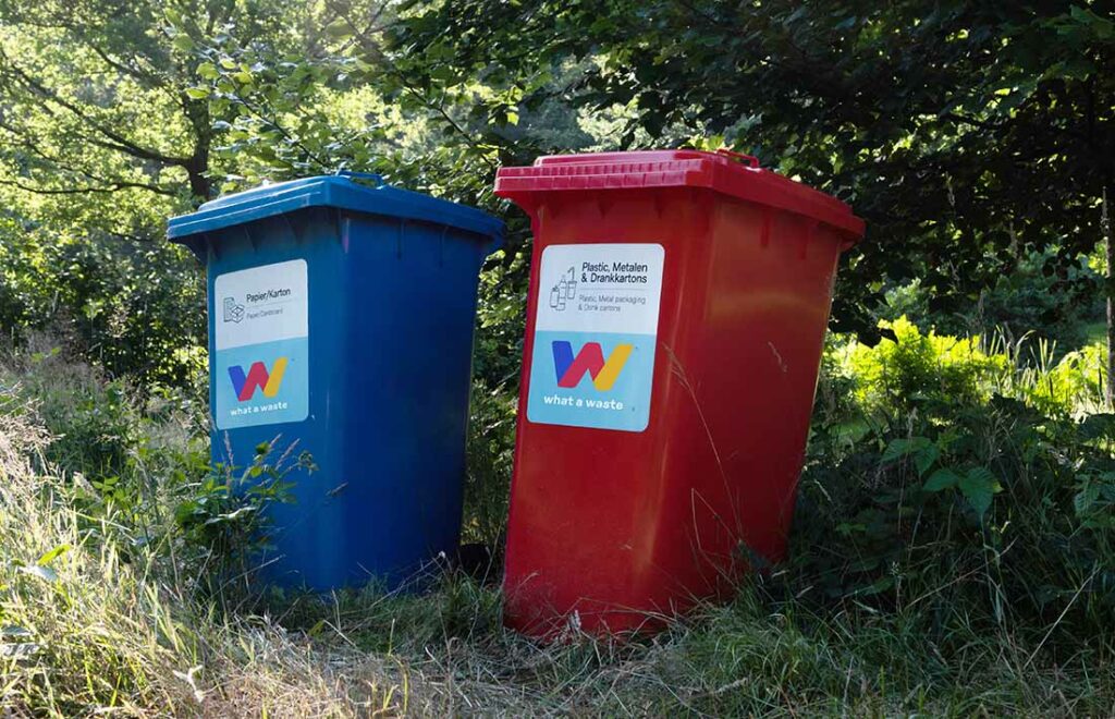 Twee rolcontainers van What A Waste, een blauwe voor papier/karton en een rode voor plastic, geplaatst in groene buitenomgeving.