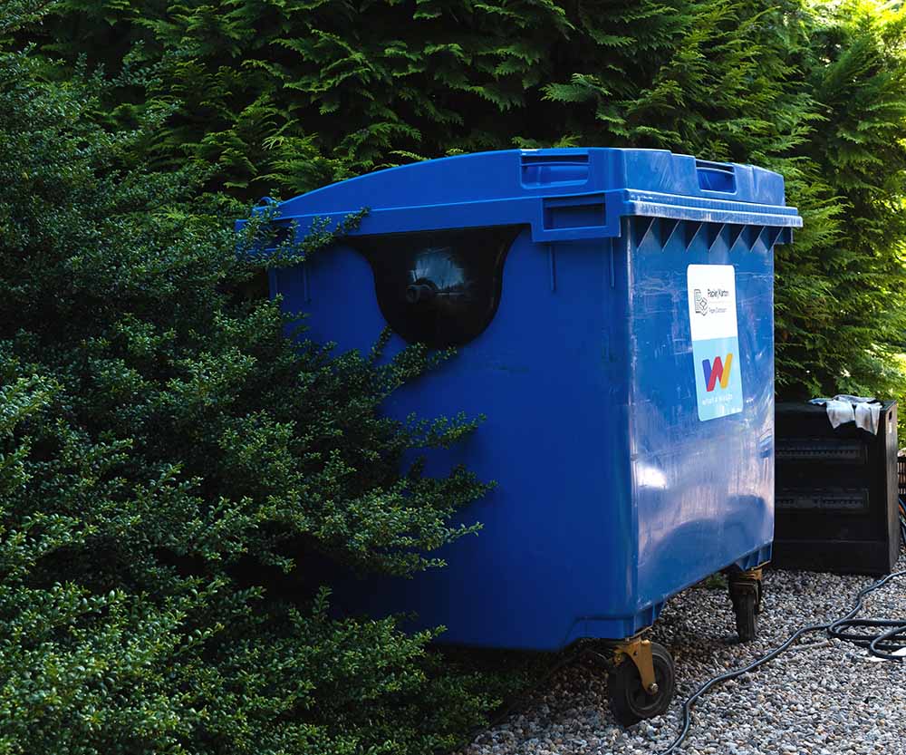 Blauwe afvalcontainer van What A Waste geplaatst in groene omgeving, geschikt voor papier/karton afvalinzameling.