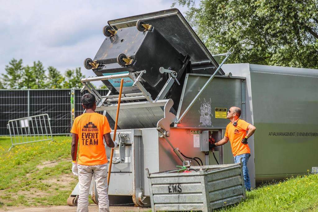 Twee medewerkers van What A Waste in oranje eventteam-shirts bedienen een perscontainer op een evenemententerrein, illustratie van duurzame afvalinzameling en containerverhuur.