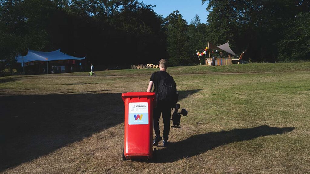 Persoon trekt een rode afvalcontainer over het grasveld bij een festivalterrein, onderdeel van de afvalbeheer werkwijze van What A Waste.